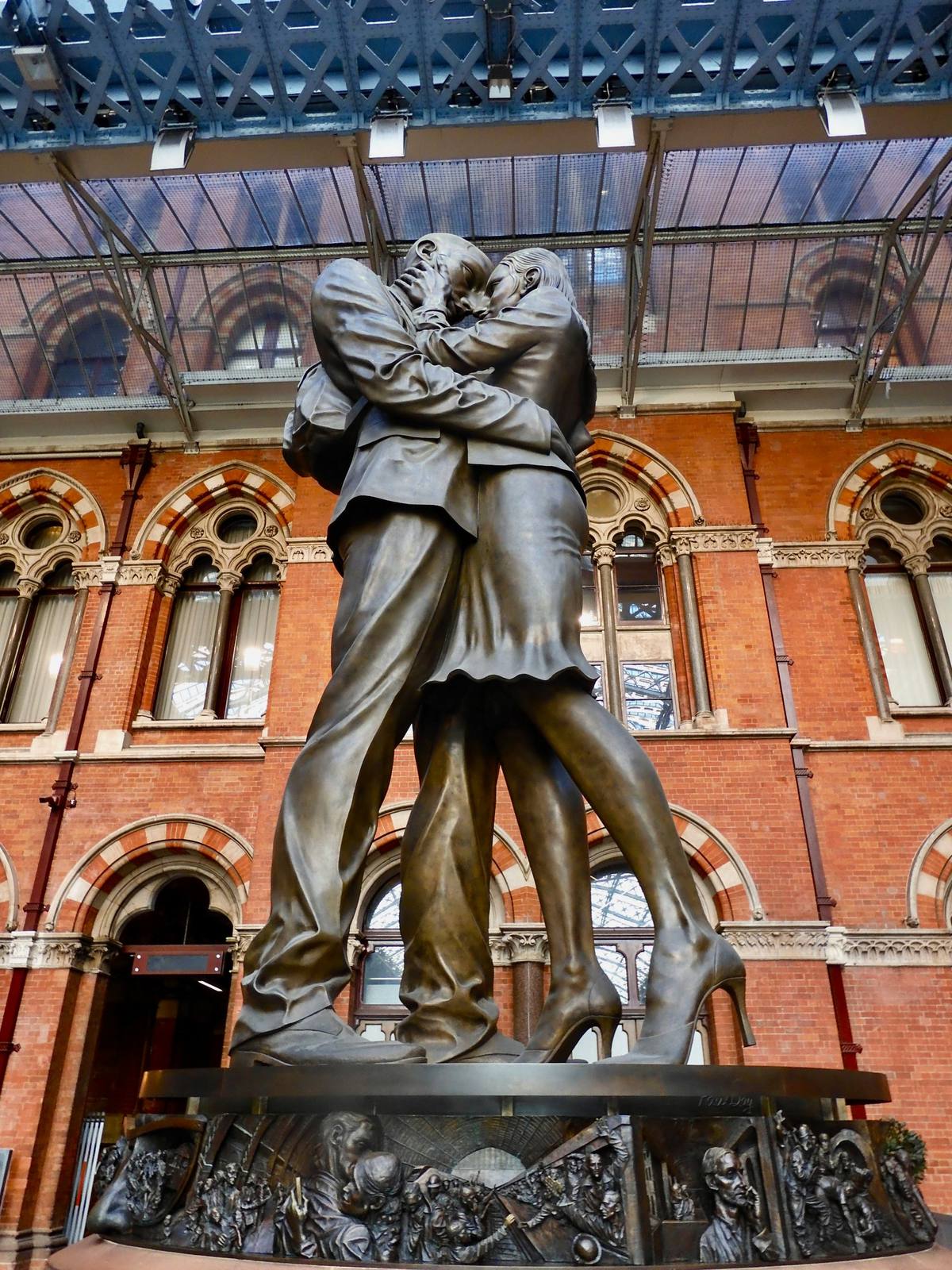 Escultura de bronce de una pareja abrazada bajo el techo de hierro de una estación, con relieves de figuras en la base