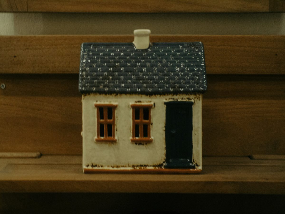 Casa de cerámica en miniatura, con tejado azul, ventanas de marco naranja y puerta oscura, sobre un estante de madera