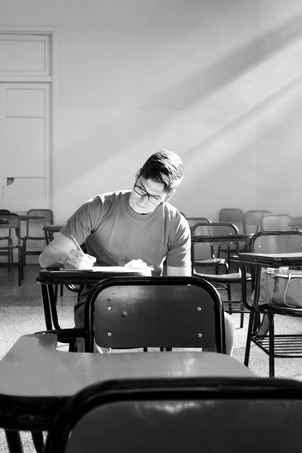 Joven escribiendo en un cuaderno sentado en un salón de clases vacío, en blanco y negro