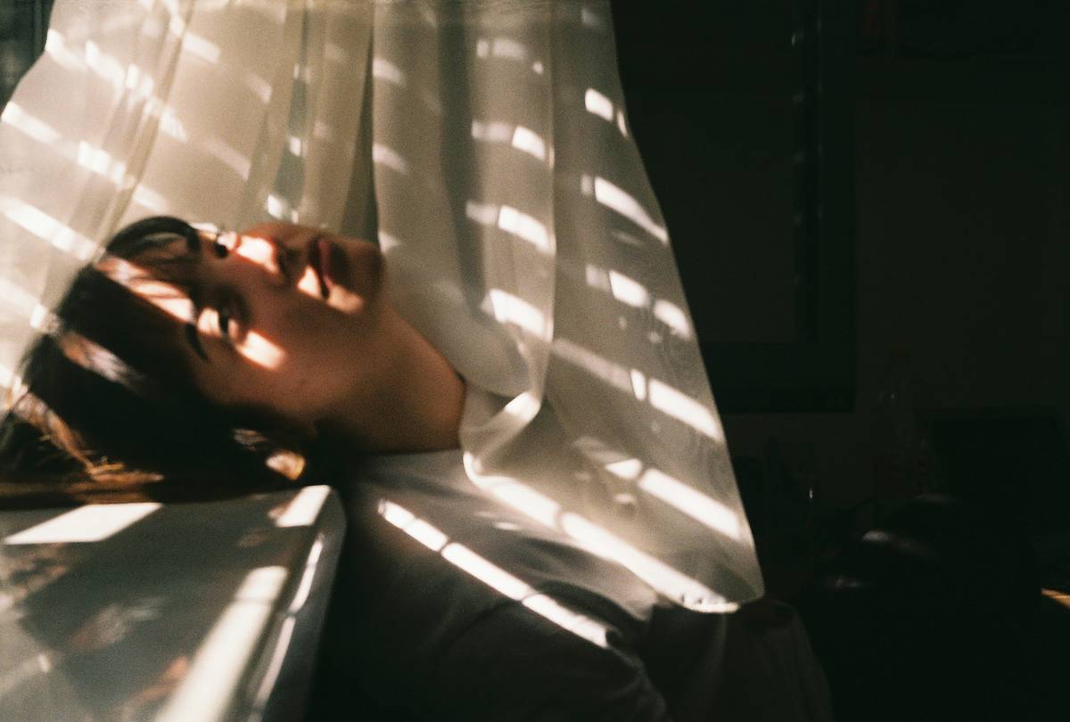 Mujer recostada entre cortinas blancas con franjas de luz solar atravesando la escena