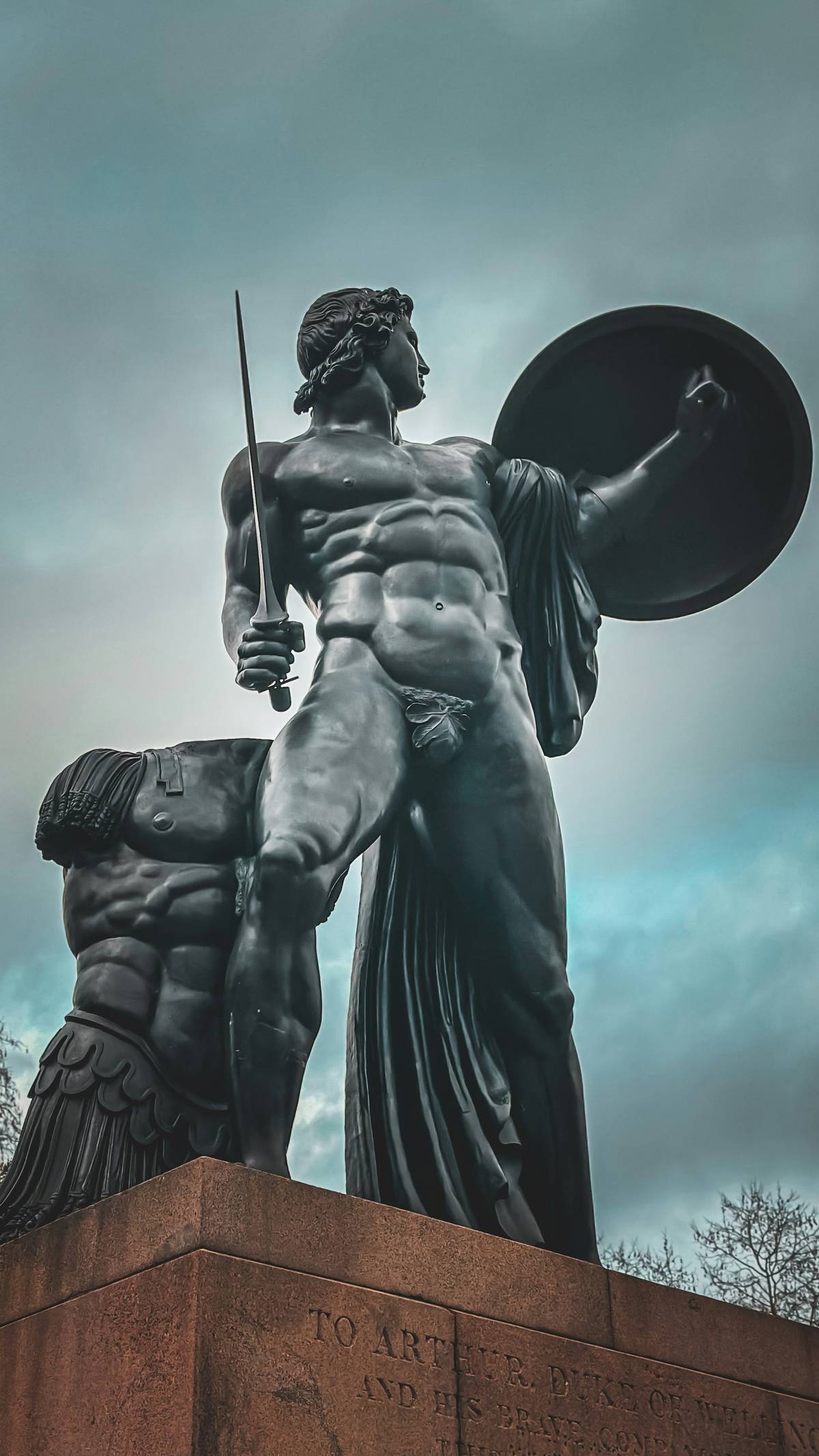 Estatua de bronce de un guerrero heroico de pie sobre un pedestal, con espada y escudo, junto a una figura caída