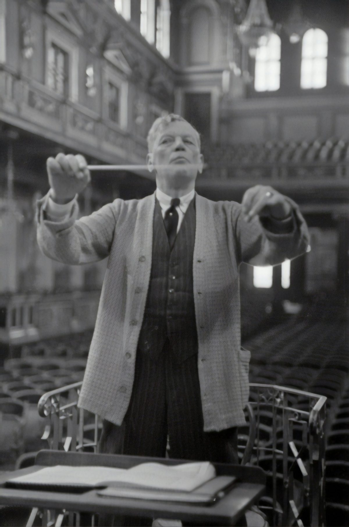 Director de orquesta con chaqueta de punto sobre chaleco y corbata, con la batuta en alto, frente a las butacas vacías de una sala, en blanco y negro
