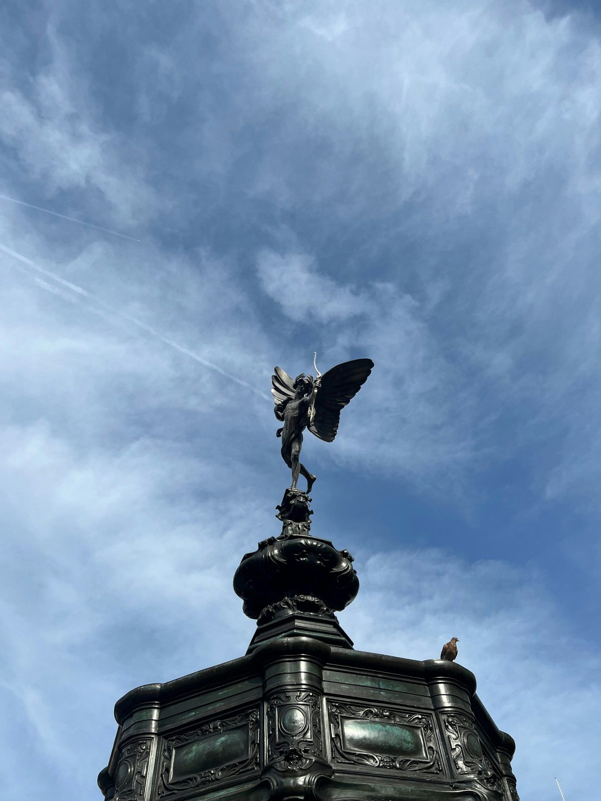Estatua de bronce alada de Anteros, con arco en mano, sobre una fuente ornamentada contra un cielo azul con nubes