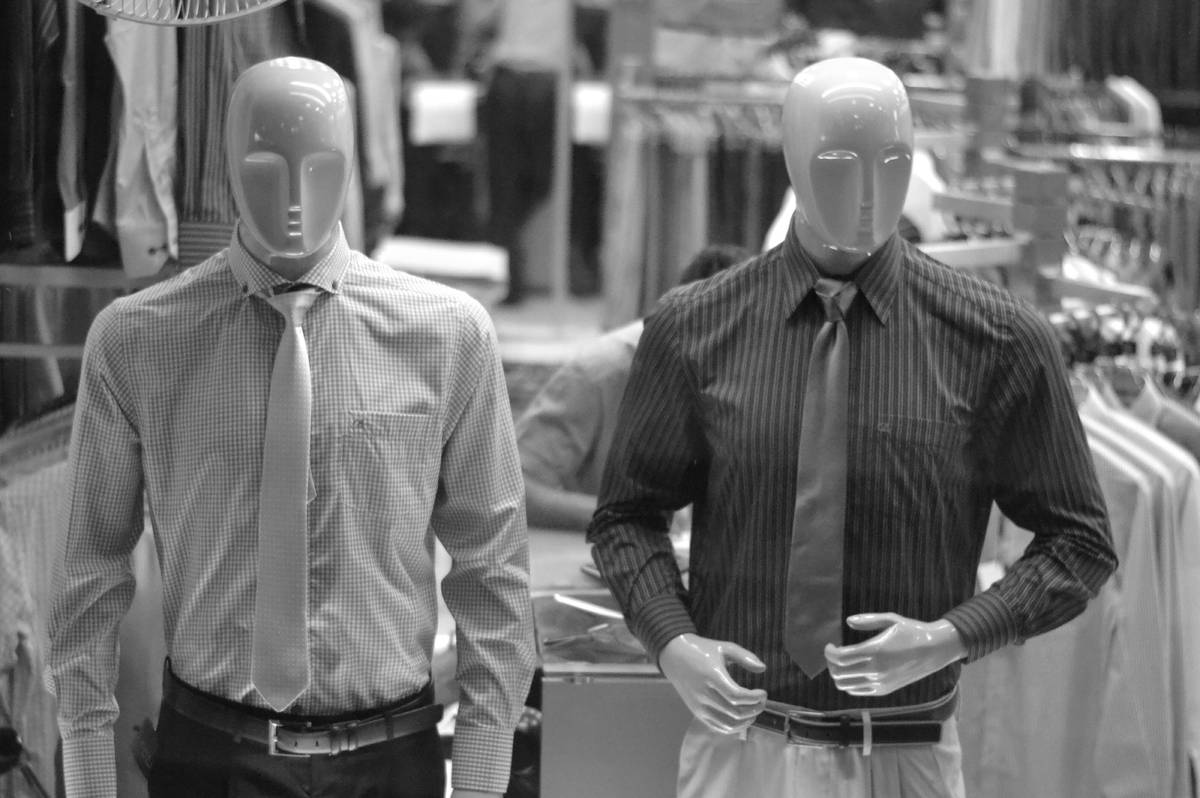 Dos maniquíes sin rasgos faciales vestidos con camisa y corbata en una tienda de ropa, en blanco y negro