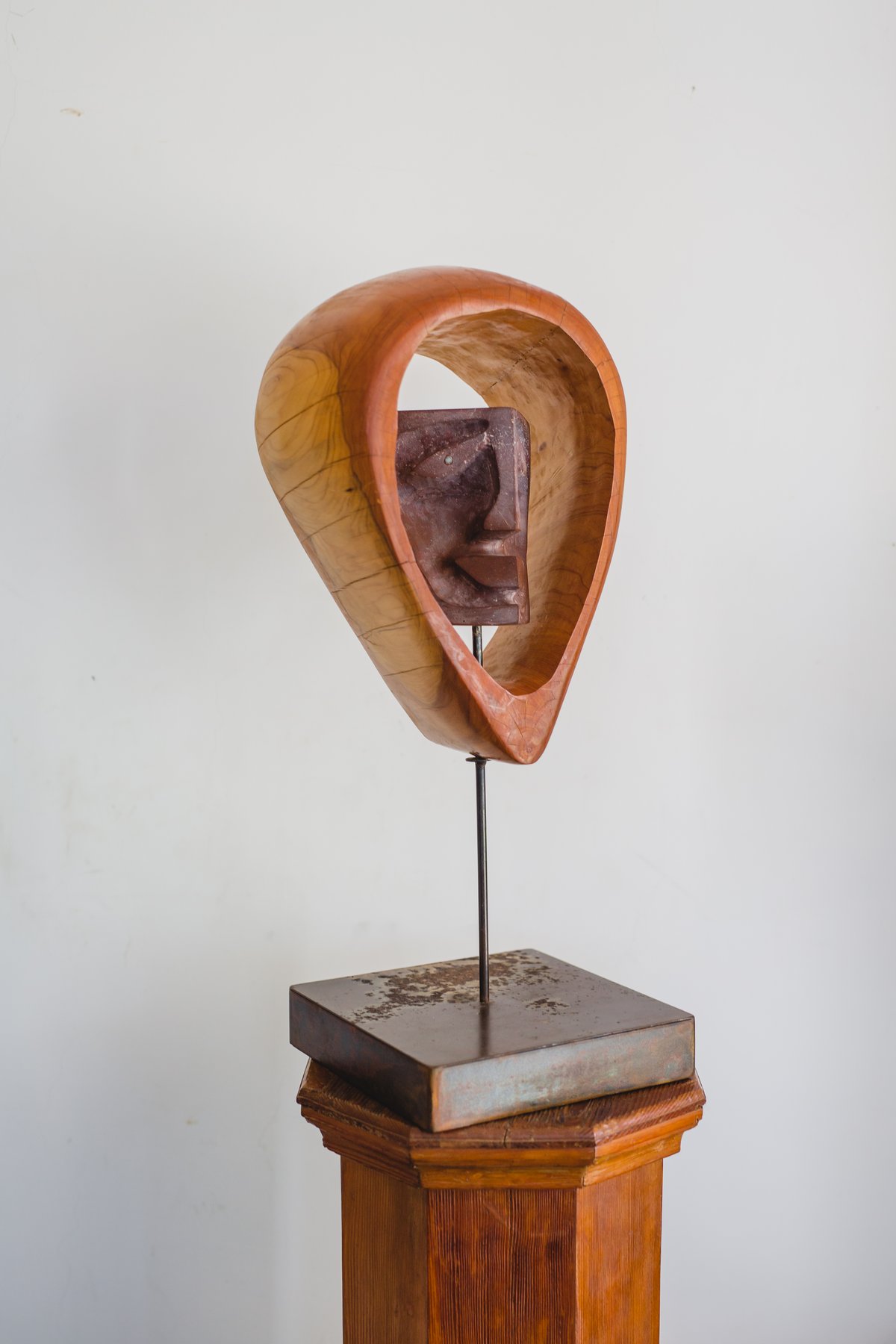 Escultura de madera curvada en forma de gota invertida con un rostro tallado en piedra oscura suspendido en su interior