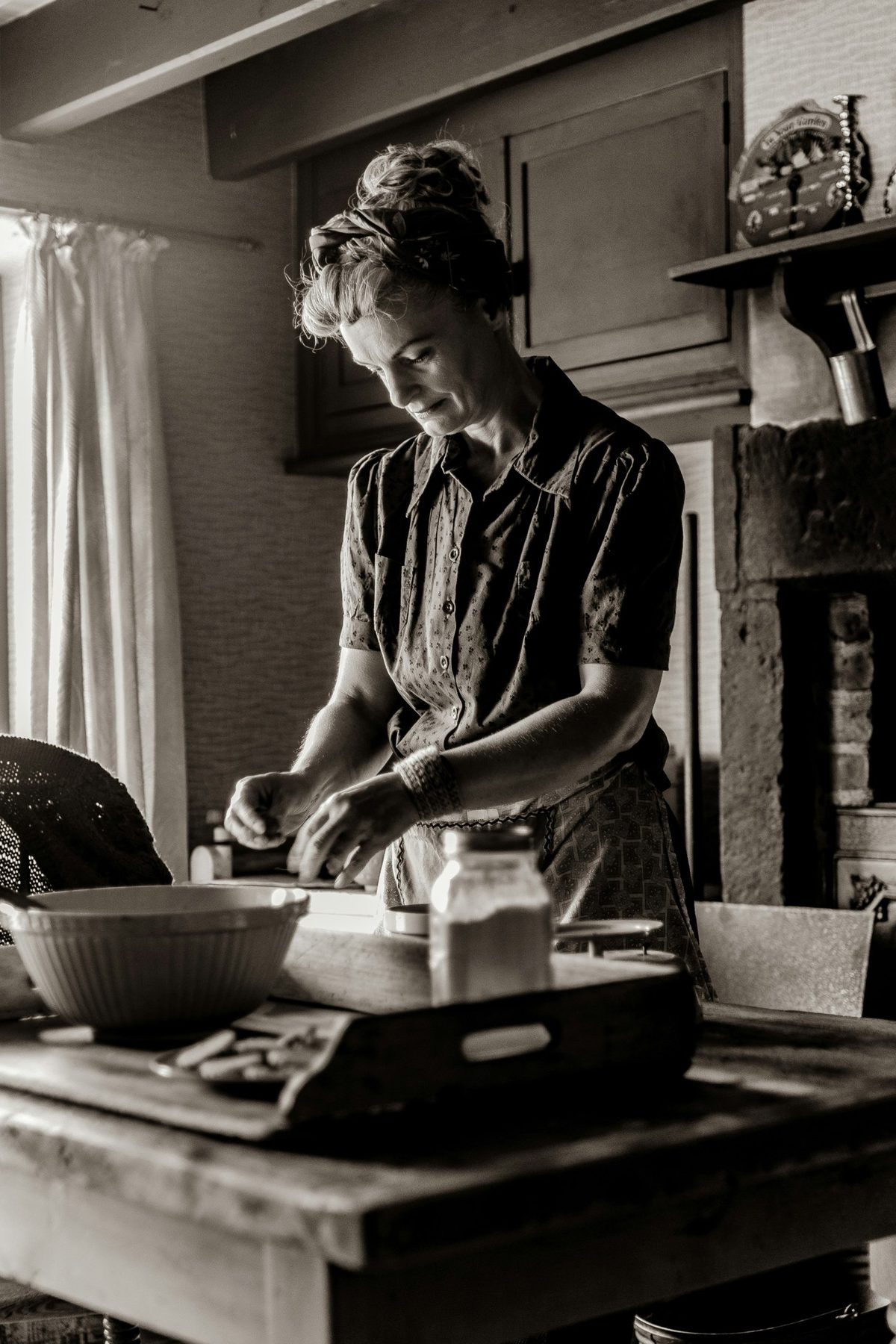 Mujer con un pañuelo en el pelo trabajando la masa sobre una mesa de madera en una cocina antigua, en sepia