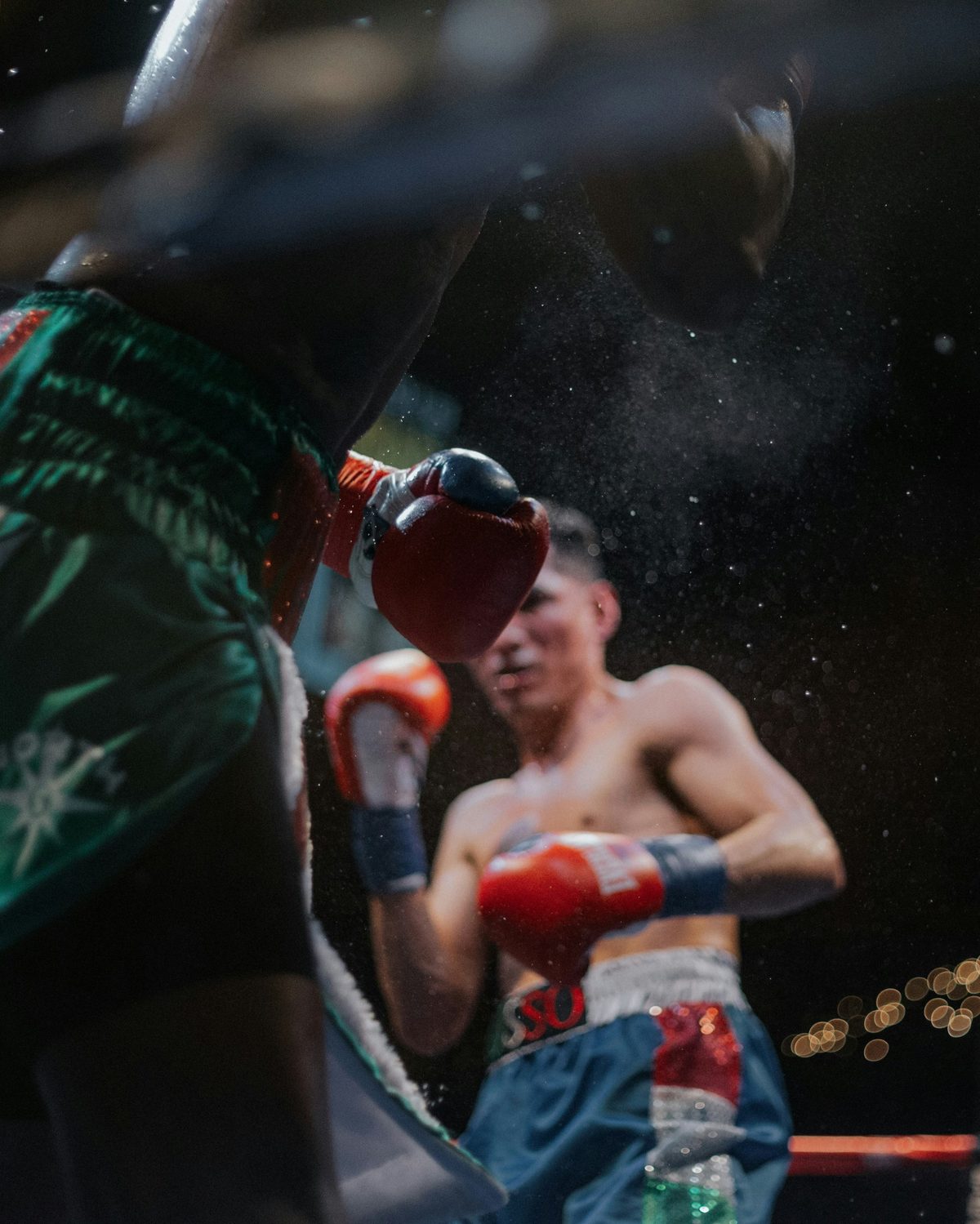 Dos boxeadores en un ring: uno desenfocado de espaldas en primer plano y otro con guantes rojos al fondo, con partículas de sudor en el aire